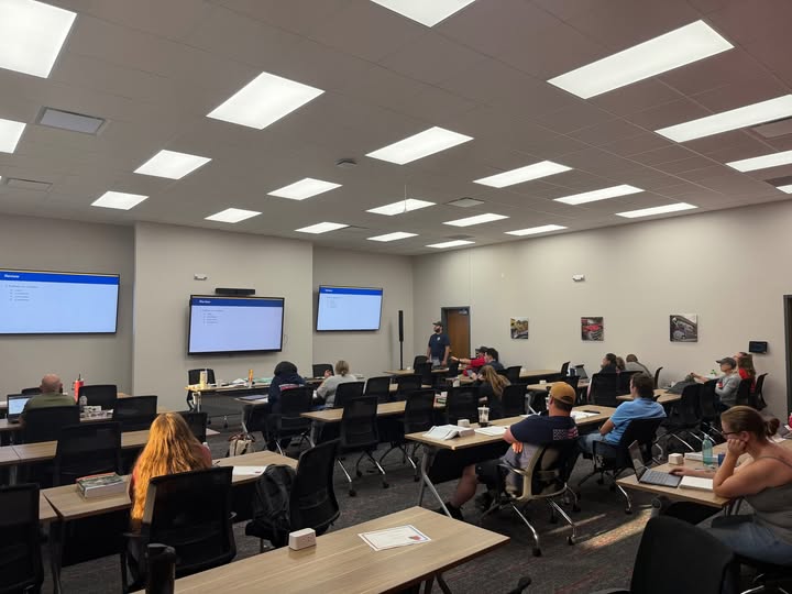 A classroom full of students sitting at tables facing three large wall-mounted screens displaying presentations.