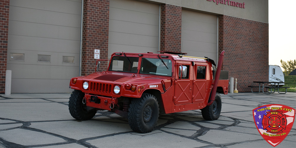 Red North Sioux City Fire Department brush truck parked in front of a fire station, with the department’s patch overlaid in the corner.