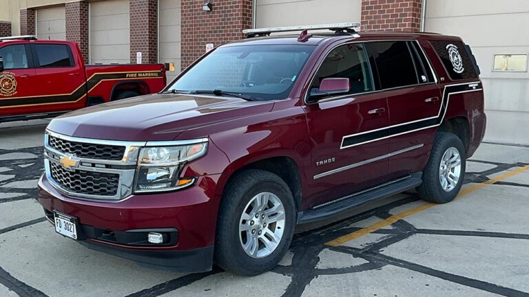 A maroon fire department Chevrolet Tahoe SUV parked in front of a brick station with a red fire marshal truck visible in the background.