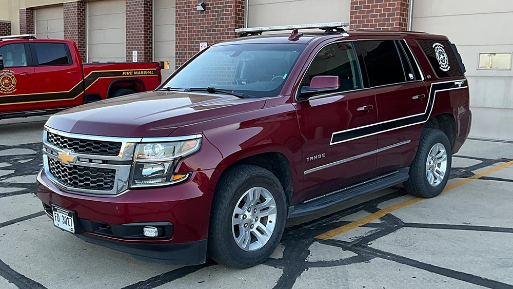 A maroon fire department Chevrolet Tahoe SUV parked in front of a brick station with a red fire marshal truck visible in the background.
