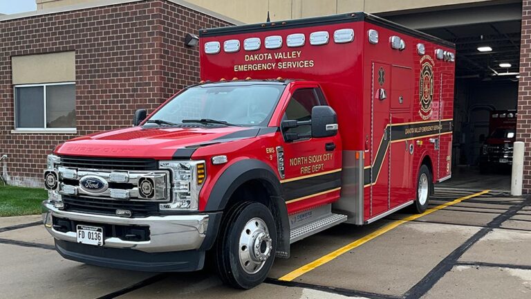 A bright red Dakota Valley Emergency Services ambulance parked outside a brick fire station.