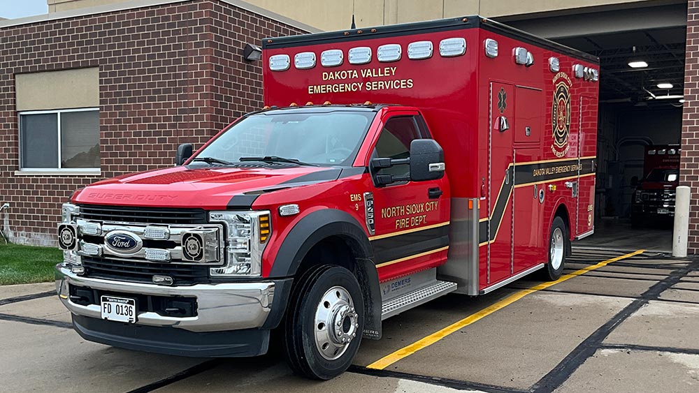 A bright red Dakota Valley Emergency Services ambulance parked outside a brick fire station.