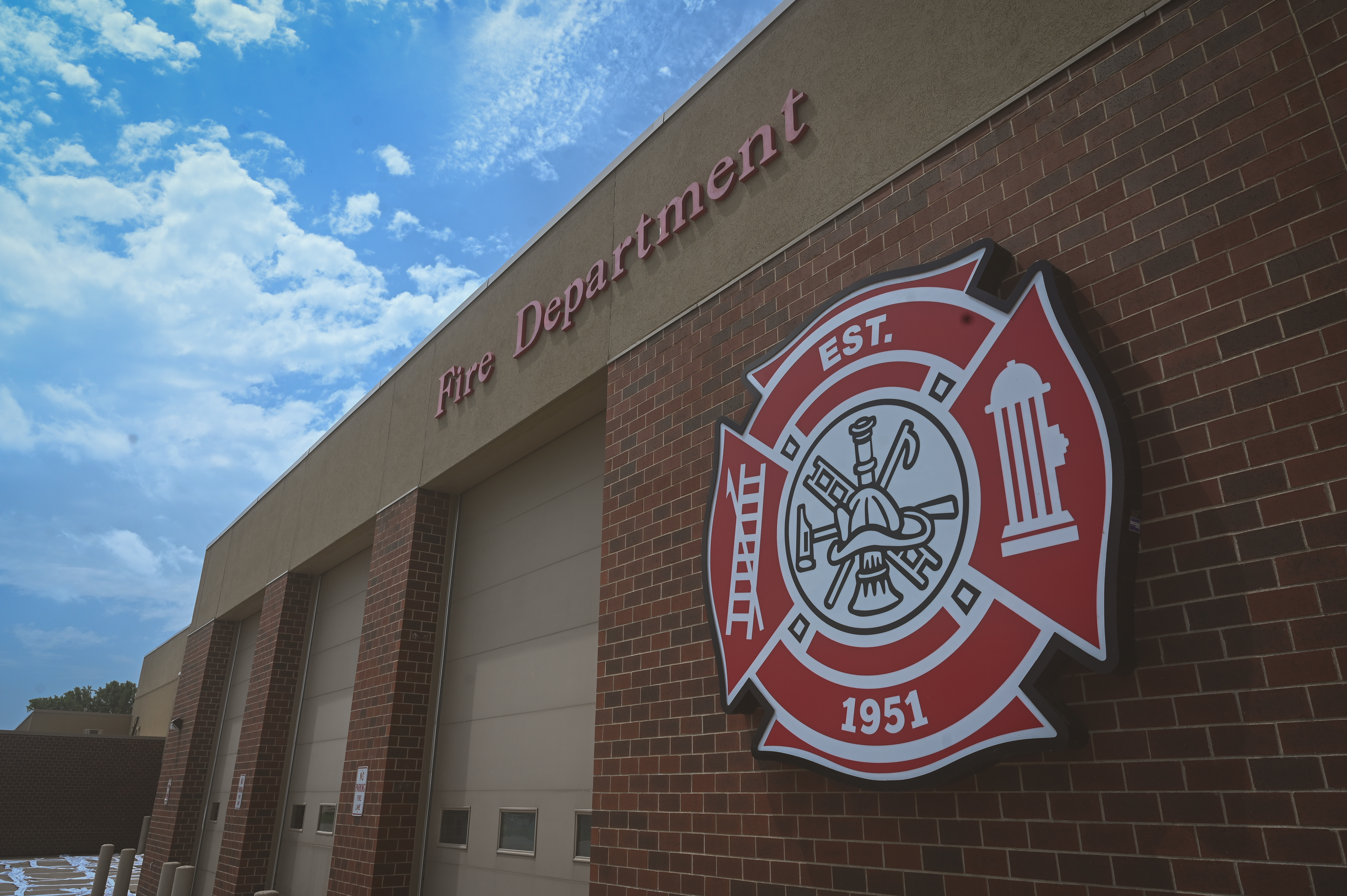The exterior of a brick fire station featuring a large red and white Maltese cross emblem that reads “EST. 1951” and the words “Fire Department” mounted above the garage doors.