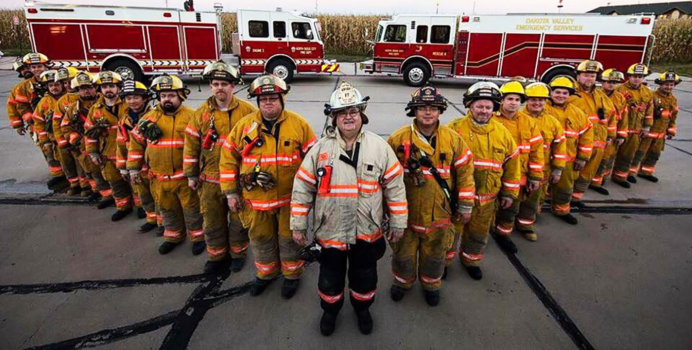 A group of firefighters in full turnout gear stand in rows in front of two parked fire trucks.