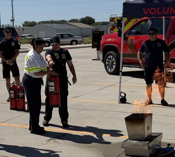A firefighter instructor assists a woman in using a fire extinguisher to put out a controlled fire in a metal training container.