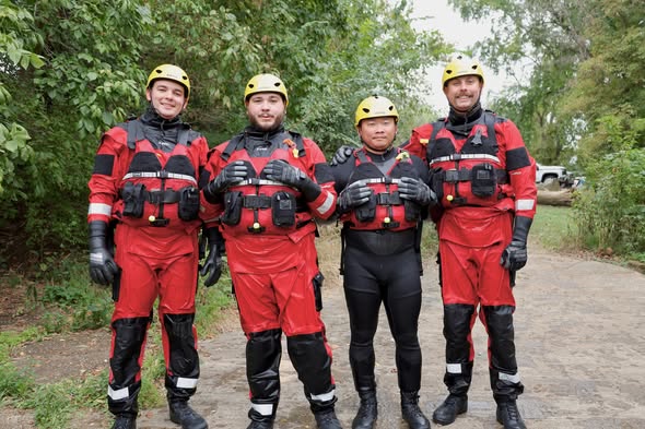 Four people in red and black rescue drysuits and yellow helmets stand together outdoors.