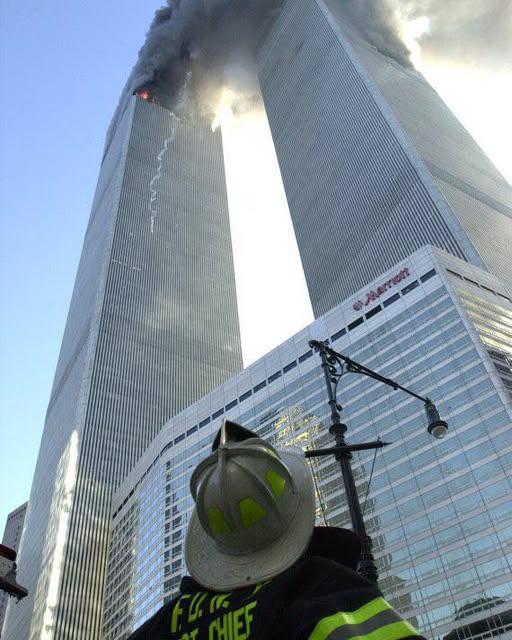 A firefighter stands in the foreground, facing the Twin Towers as smoke billows from the upper floors.