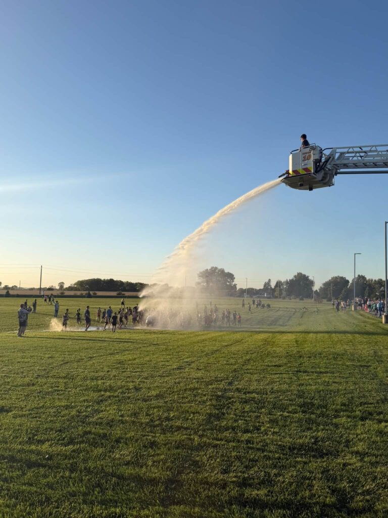 A fire truck ladder sprays water over a large group of people running across a grassy field.
