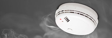 White smoke detector mounted on a ceiling surrounded by wisps of smoke.