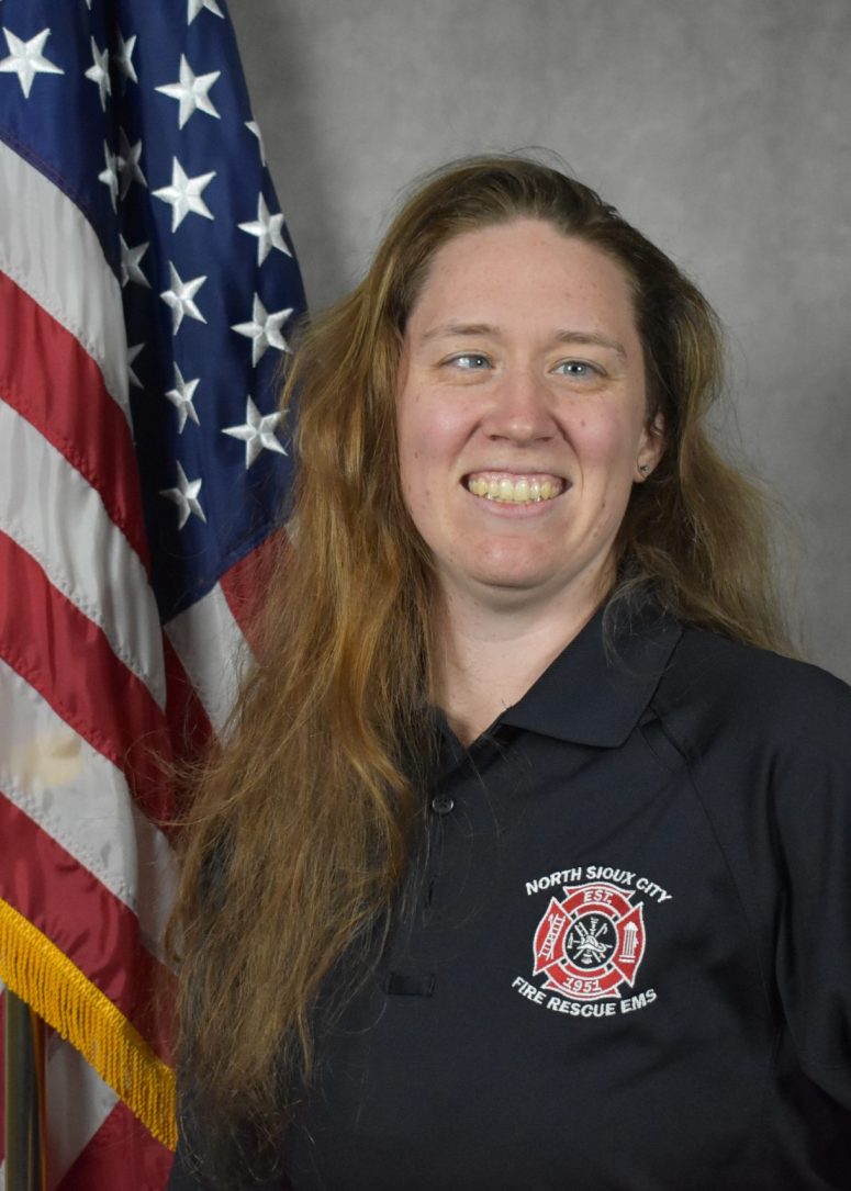 A smiling woman in a North Sioux City Fire Rescue EMS polo shirt stands in front of an American flag.