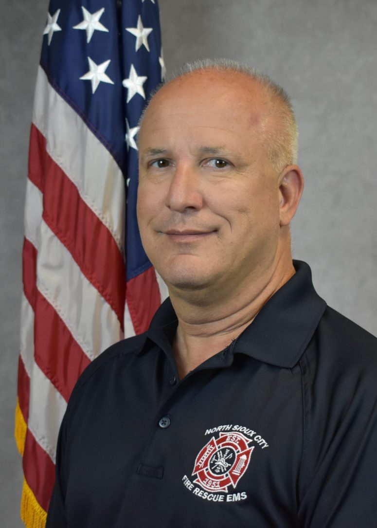 A man in a navy blue North Sioux City Fire Rescue EMS polo shirt stands in front of an American flag.