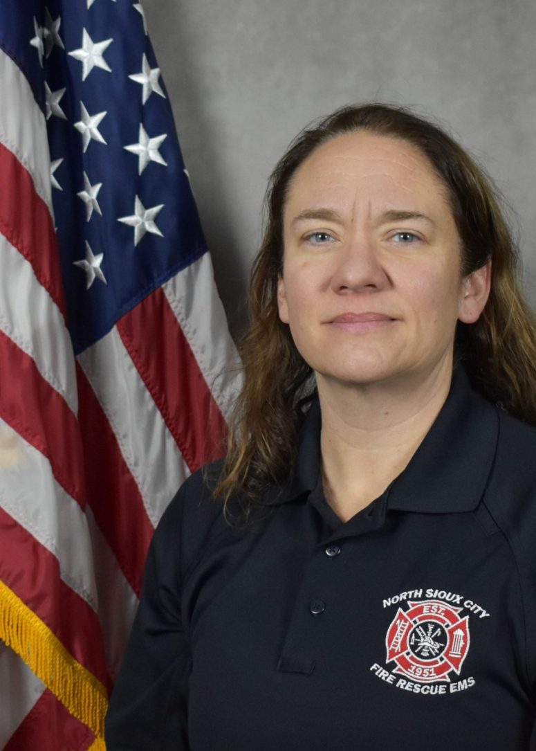A North Sioux City Fire Rescue EMS official in a navy polo shirt stands in front of an American flag.