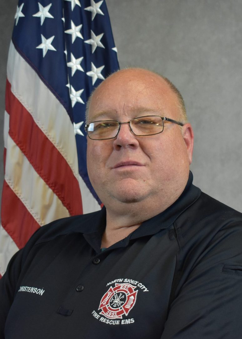 A man in a black North Sioux City Fire Rescue EMS polo shirt standing in front of an American flag.
