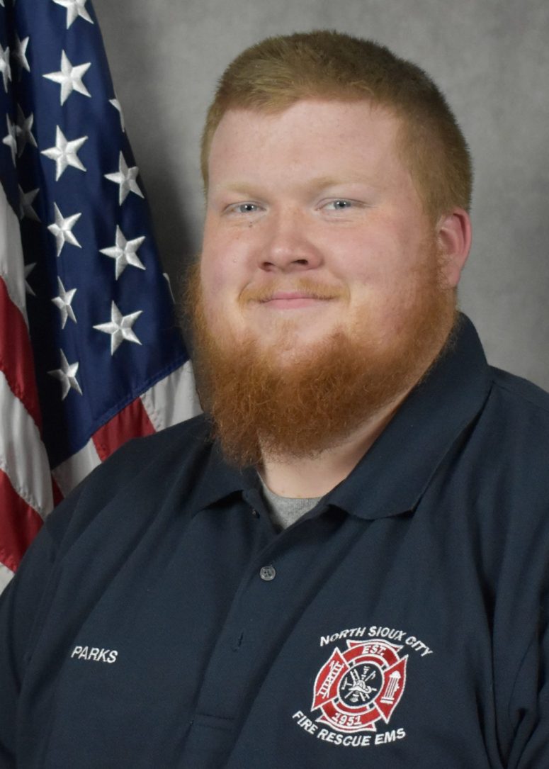 A North Sioux City Fire Rescue EMS official in a navy polo shirt stands in front of an American flag.