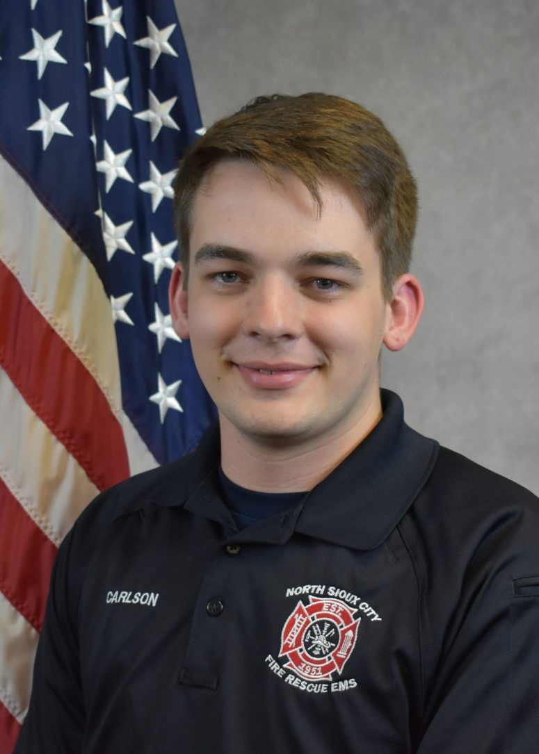 A young man in a North Sioux City Fire Rescue EMS polo shirt stands in front of an American flag.