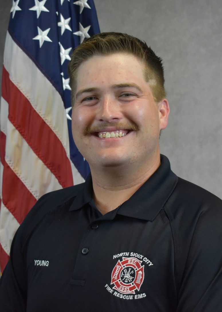 A smiling firefighter in a dark uniform with a North Sioux City Fire Rescue EMS patch, standing in front of an American flag.