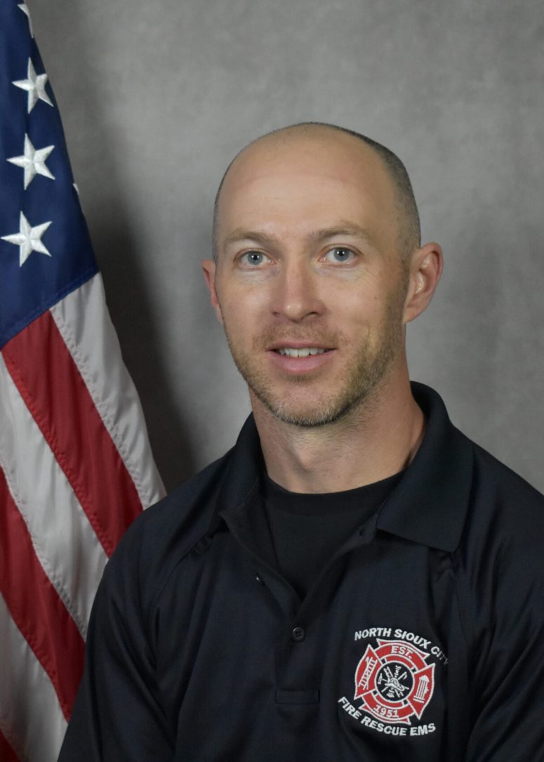 A firefighter in a black uniform standing in front of an American flag.