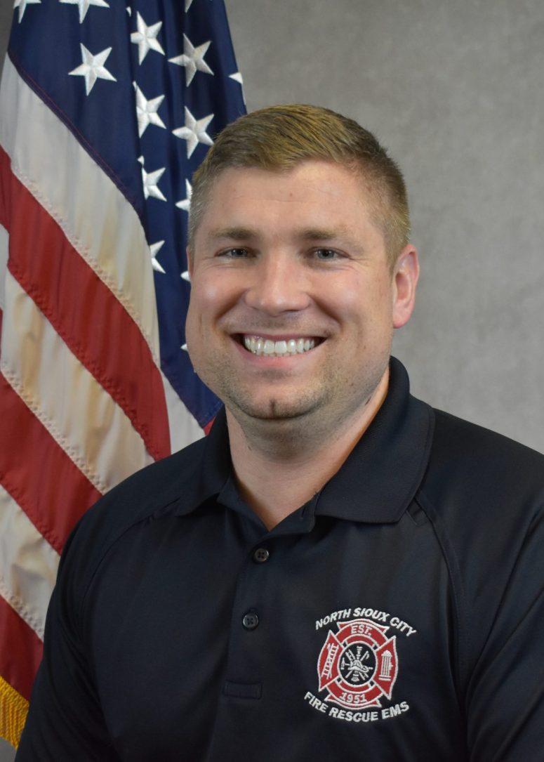 A smiling firefighter in a black uniform shirt with a North Sioux City Fire Rescue EMS emblem, standing in front of an American flag.