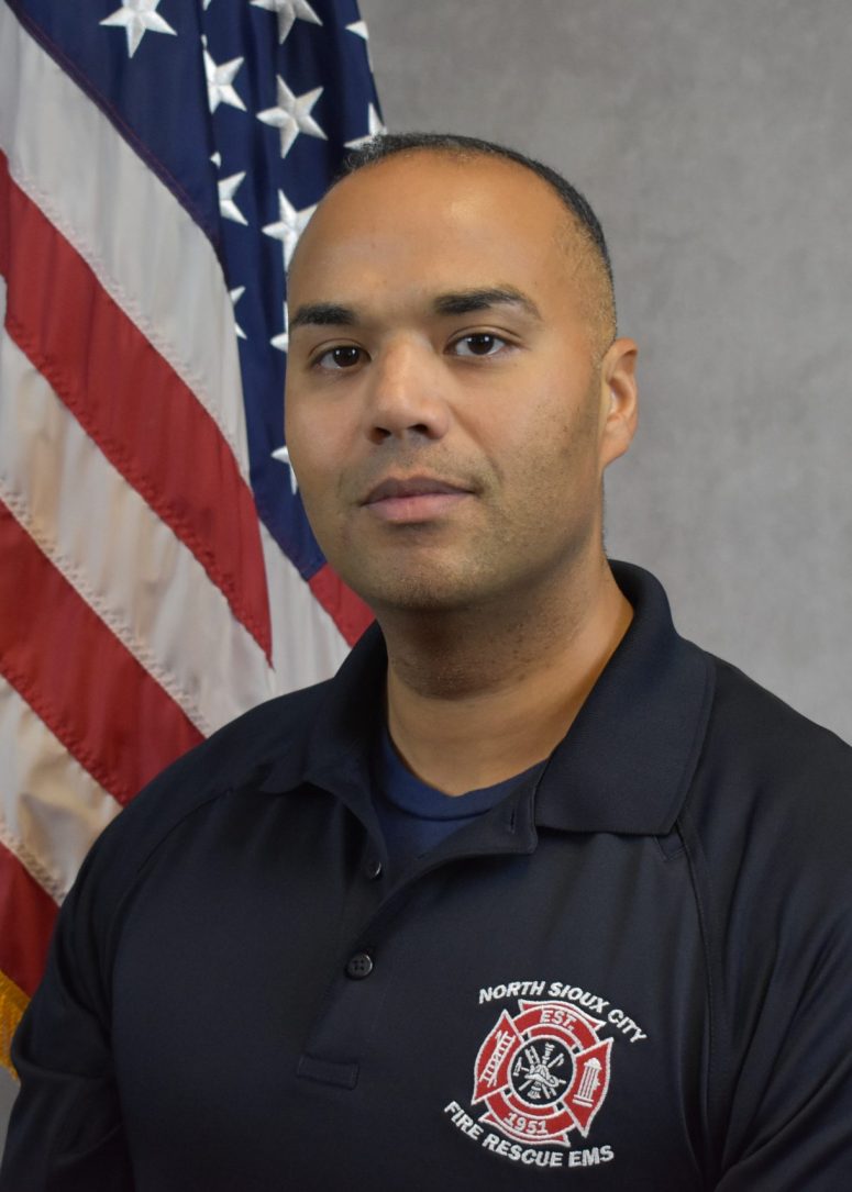A North Sioux City Fire Rescue EMS official in a navy blue uniform stands in front of an American flag.