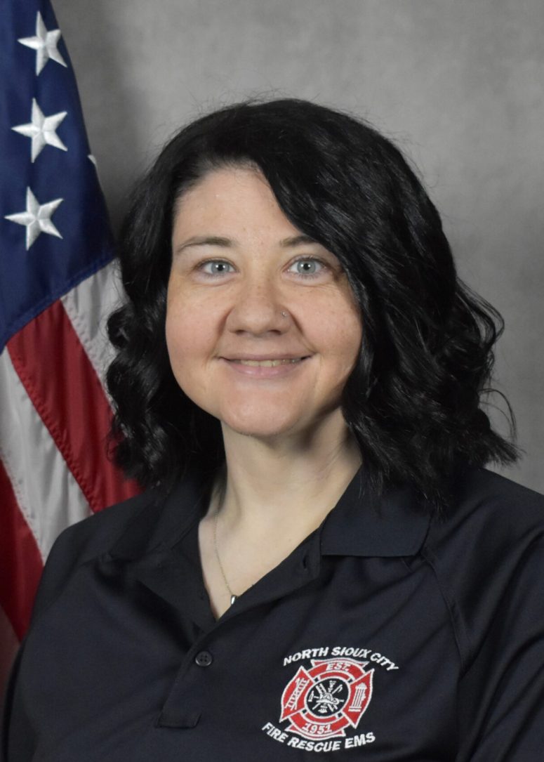 A woman wearing a North Sioux City Fire Rescue EMS polo shirt stands in front of an American flag.