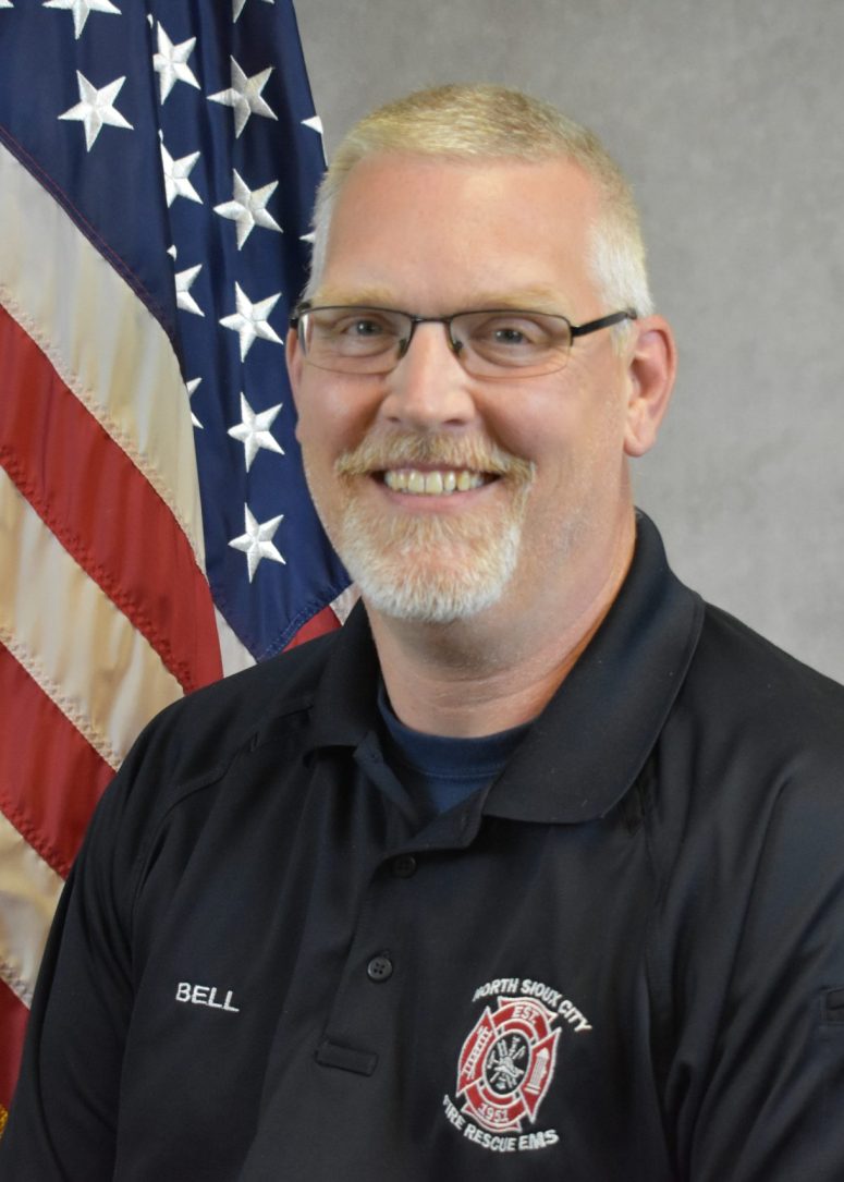 A smiling firefighter wearing a black uniform shirt with a North Sioux City Fire Rescue patch and a name tag reading “BELL,” standing in front of an American flag.
