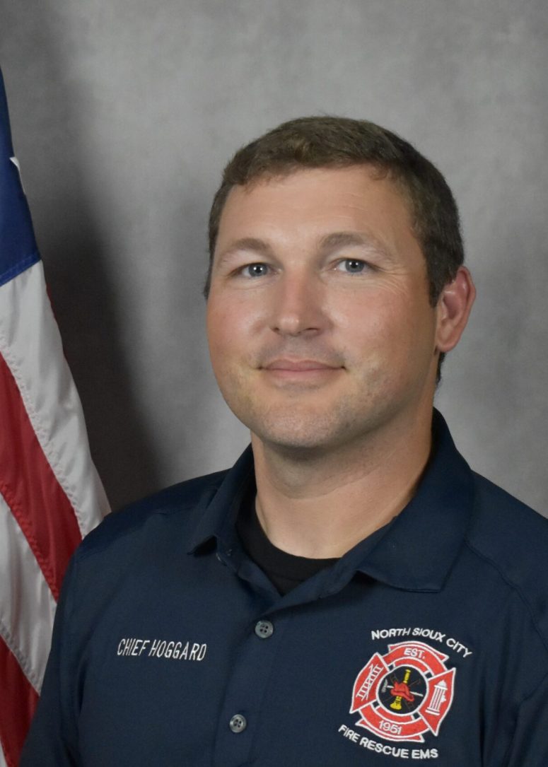 A man in a navy blue North Sioux City Fire Rescue uniform standing in front of an American flag.