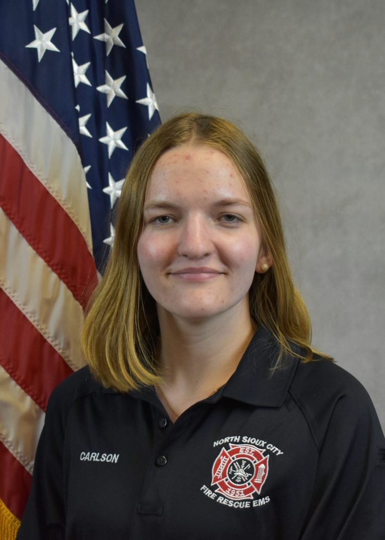 A young woman wearing a black North Sioux City Fire Rescue EMS polo shirt stands in front of an American flag.