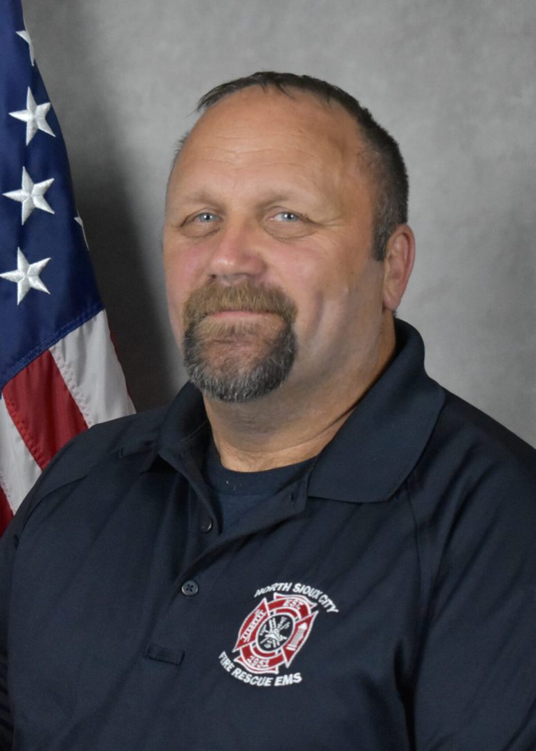 A middle-aged man in a dark polo shirt with a North Sioux City Fire Rescue EMS patch, standing in front of an American flag.
