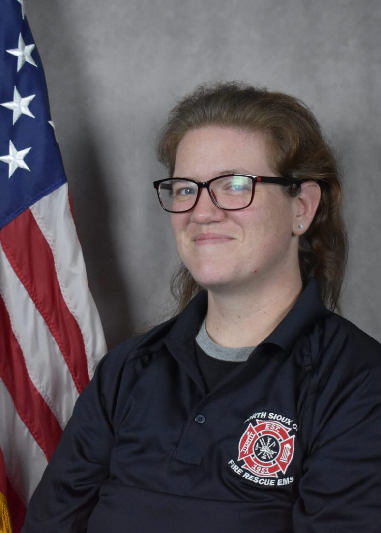 A person wearing glasses and a North Sioux City Fire Rescue EMS polo shirt poses in front of an American flag.