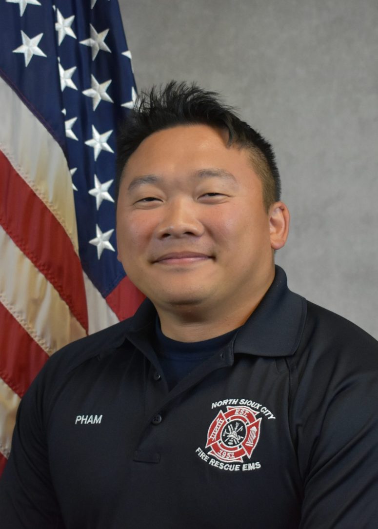 A North Sioux City Fire Rescue EMS firefighter in a uniform polo shirt stands in front of an American flag.
