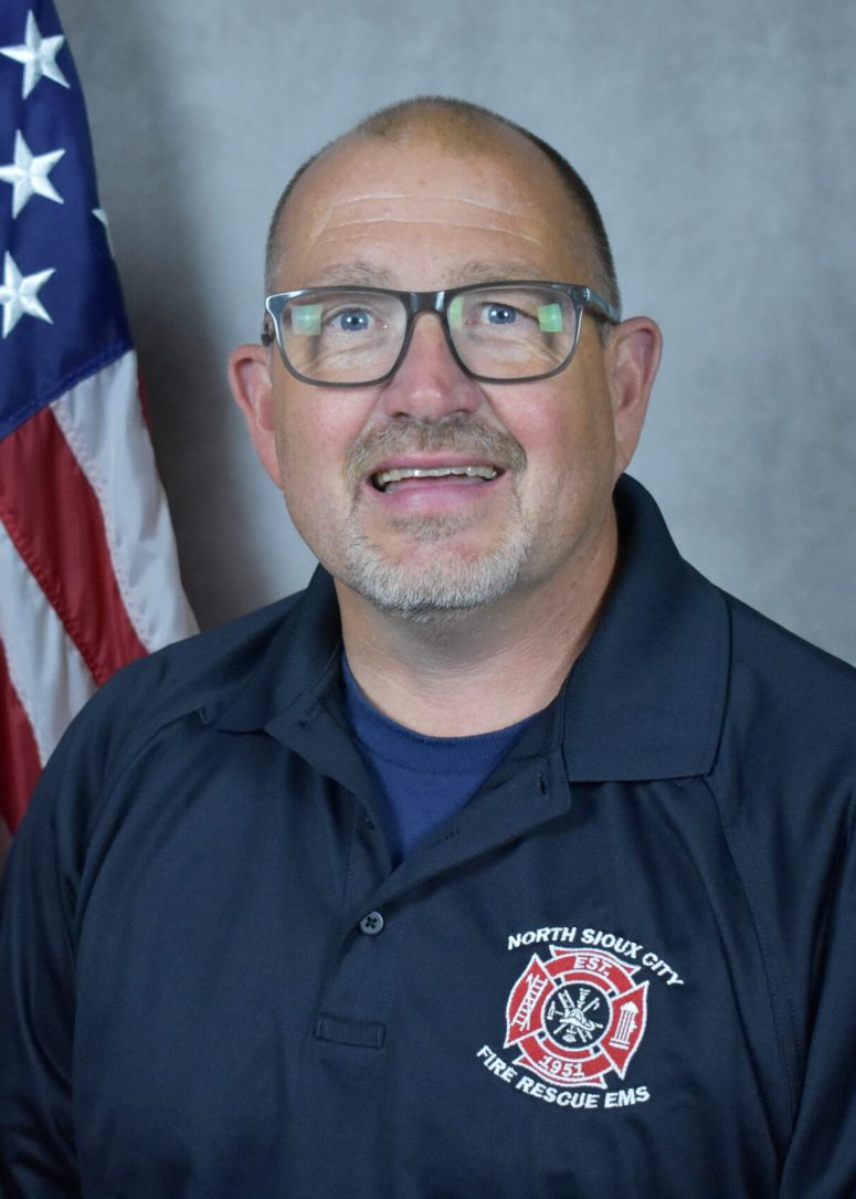 A middle-aged man wearing glasses and a North Sioux City Fire Rescue EMS polo shirt stands in front of an American flag.