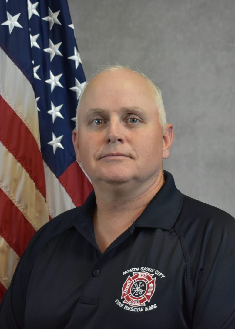 A North Sioux City Fire Rescue EMS firefighter in a navy polo shirt standing in front of an American flag.
