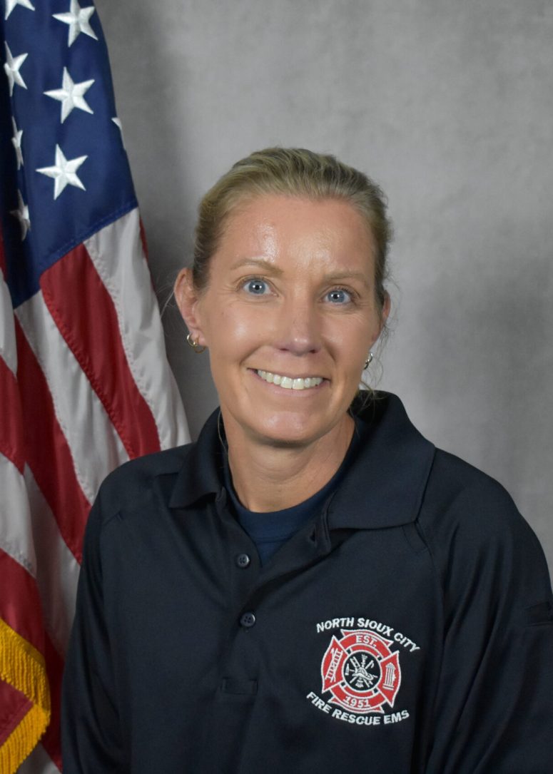 A smiling woman wearing a North Sioux City Fire Rescue EMS polo shirt, standing in front of an American flag.