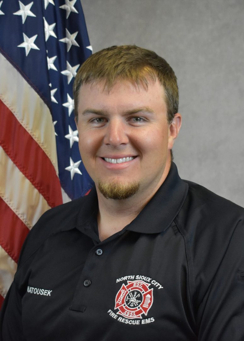 A firefighter in a black polo shirt with a North Sioux City Fire Rescue EMS patch, standing in front of an American flag.