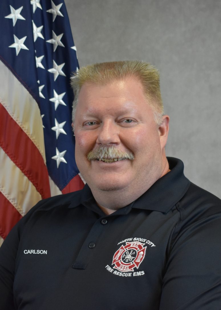 A North Sioux City Fire Rescue EMS official wearing a black polo shirt stands in front of an American flag.