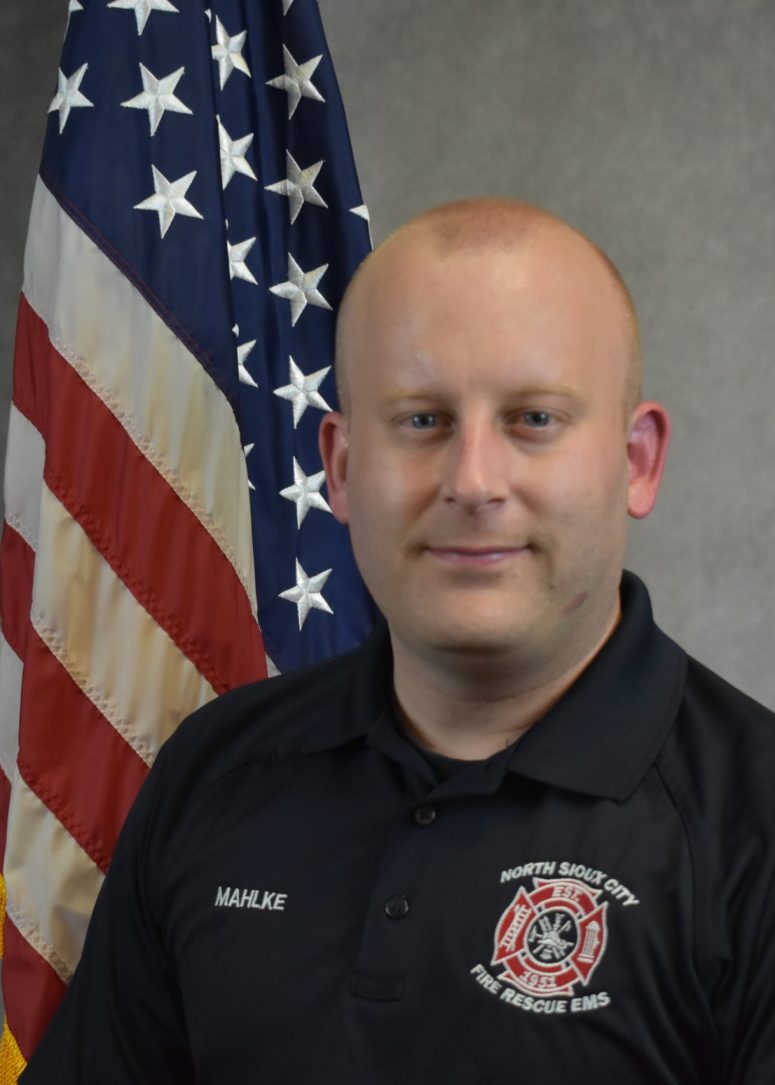 A North Sioux City Fire Rescue EMS firefighter in a black polo shirt stands in front of an American flag.