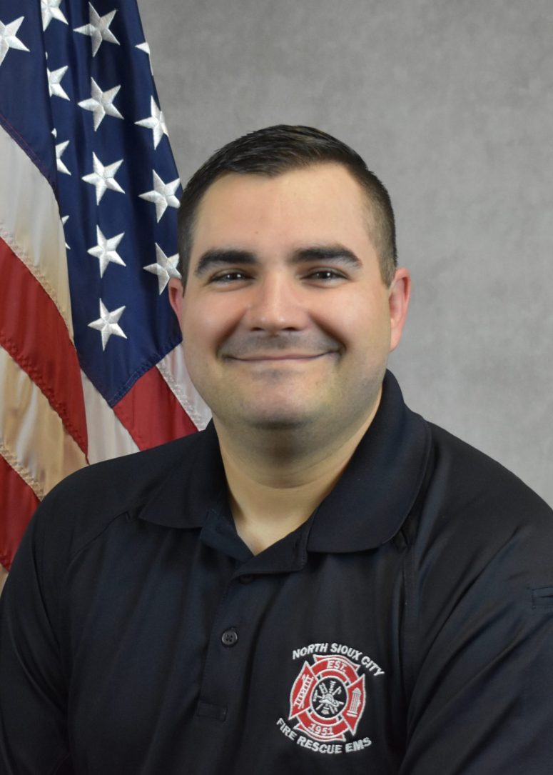 A man in a black polo shirt with a North Sioux City Fire Rescue EMS patch, standing in front of an American flag.