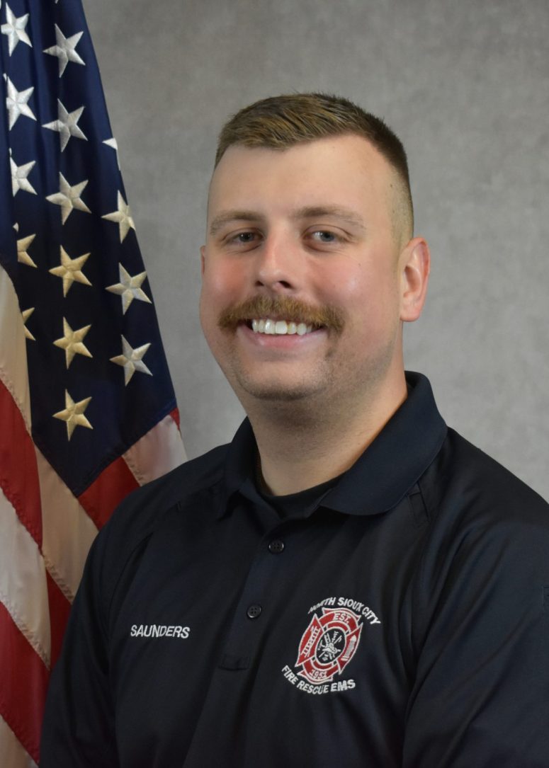 A male firefighter with a mustache wearing a dark polo shirt with a North Sioux City Fire Rescue EMS patch, standing in front of an American flag.