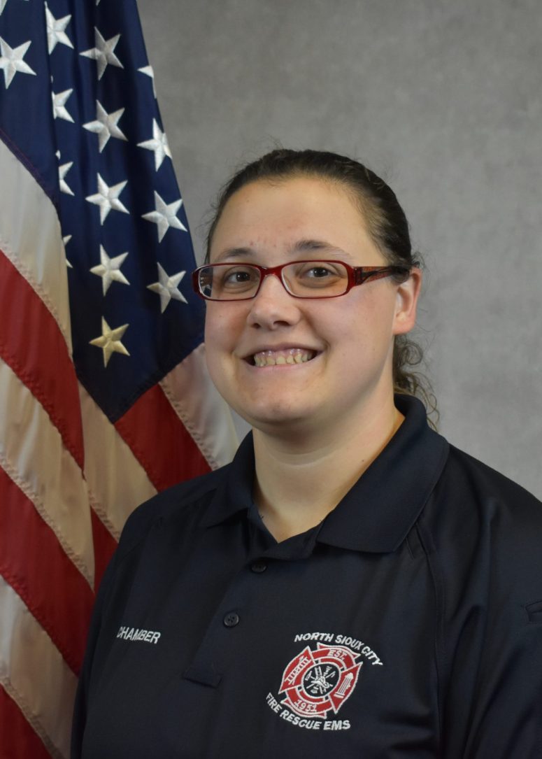 A North Sioux City Fire Rescue EMS member wearing a uniform polo shirt stands in front of an American flag.