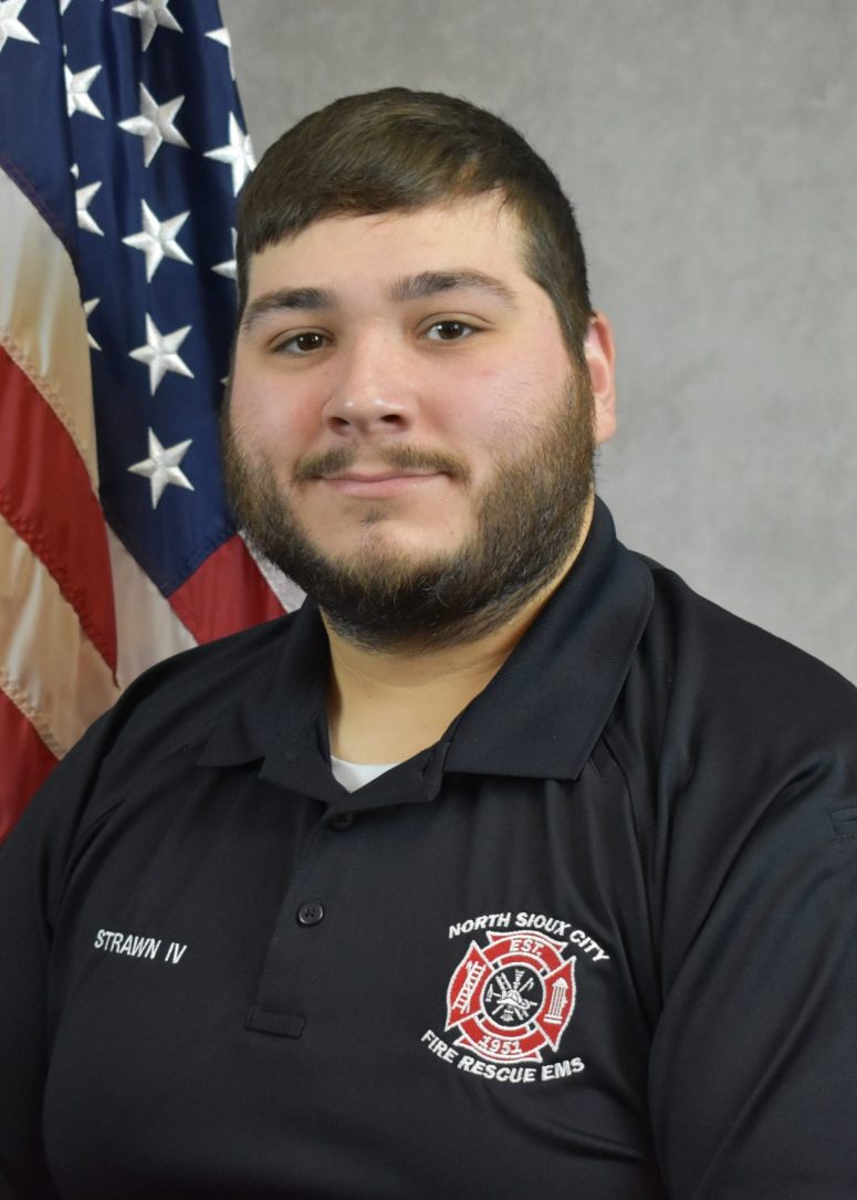 A North Sioux City Fire Rescue EMS official in a black polo shirt stands in front of an American flag.