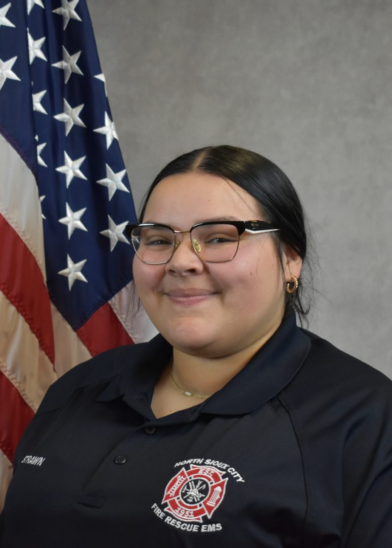 A woman wearing a dark polo shirt with a North Sioux City Fire Rescue EMS patch stands in front of an American flag.