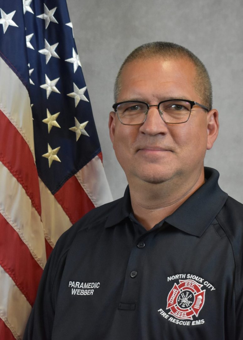 A male paramedic in a navy uniform with a North Sioux City Fire Rescue patch standing in front of an American flag.