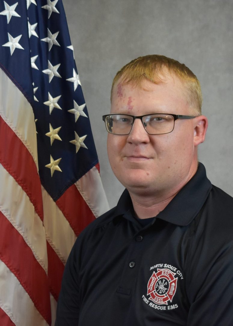A male firefighter wearing a black North Sioux City Fire Rescue EMS polo shirt, standing in front of an American flag.