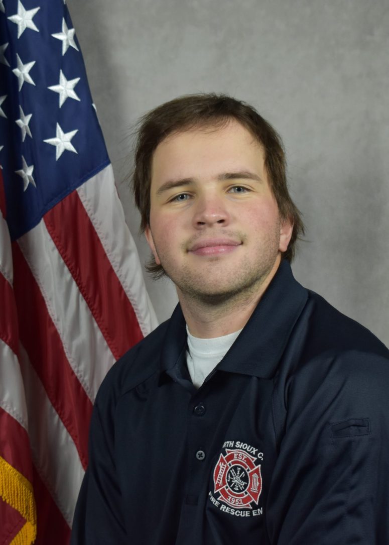 A young man in a dark blue fire department polo shirt stands in front of an American flag.