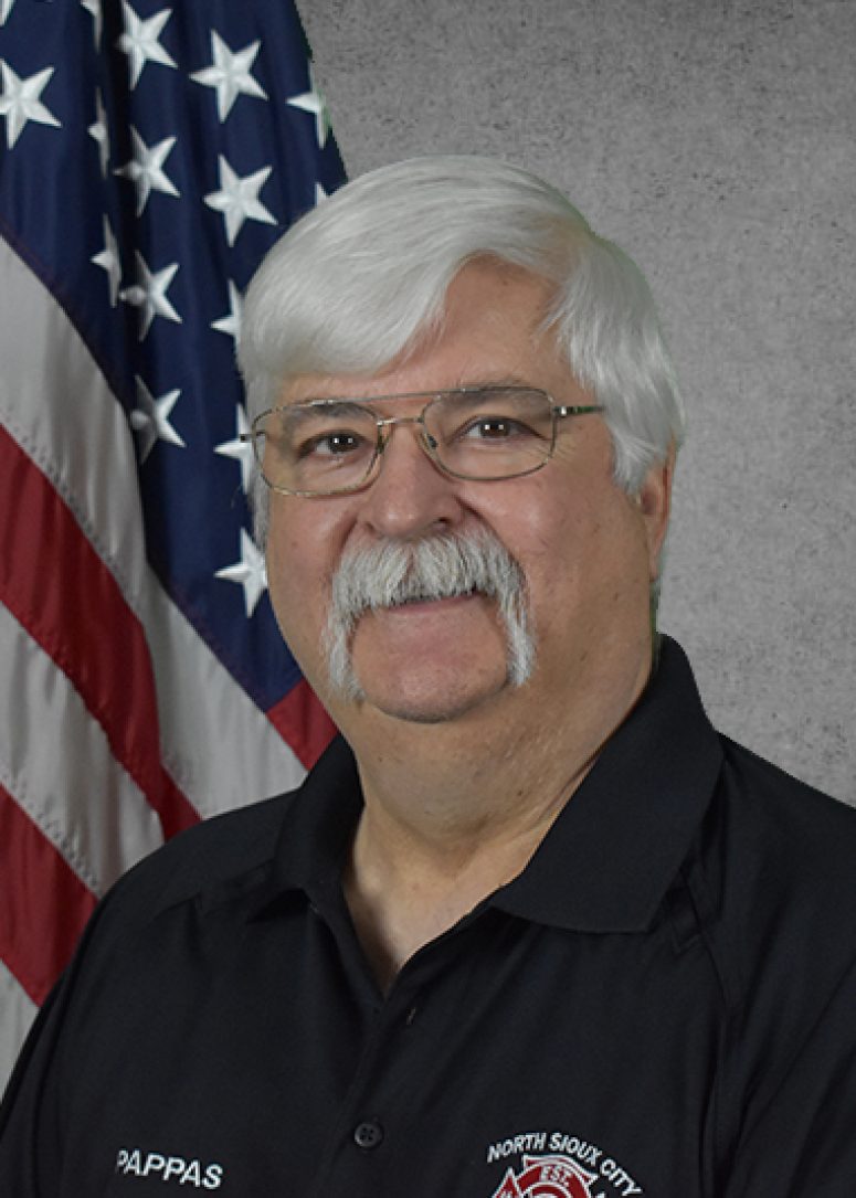 A man with a white mustache and glasses wearing a black shirt in front of an American flag.